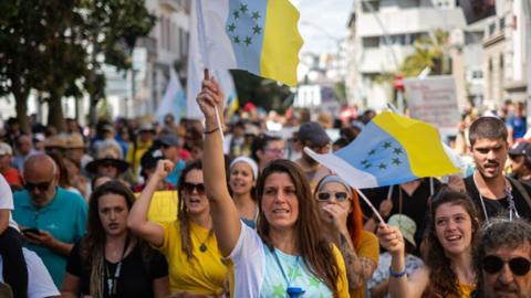 Thousands of people protesting against the tourism model and mass tourism in the Canary Islands in Tenefire, Spain on 18 May 2025. A woman in the crowd can be seen raising the flag of the Canary Islands above her head