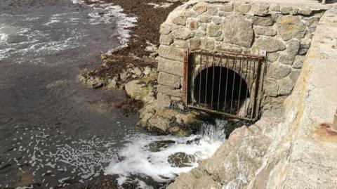 Storm overflow pipe at a beach in Cornwall. The pipe is coming from beneath a platform overlooking the sea. The hole is covered by a metal grate and liquid is pouring out of the pipe and into the sea