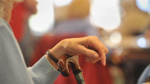 A stock image showing an elderly woman in a light blue jumper resting her arm on a walking stick with a varnished wooden handle. 