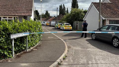 A street closed off by blue and white police tape. There is a grey sky with a police officer walking away behind the tape.
