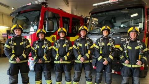 Six firefighters in full gear — helmets, jackets, pants, and boots — stand in a row inside a fire station. They face the camera with hands clasped in front. Behind them are two fire appliances, one red and one black with yellow stripes. The station’s interior, including ceiling lights, is visible in the background.