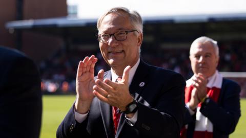 Nick Semaca standing on the pitch while clapping his hands and smiling