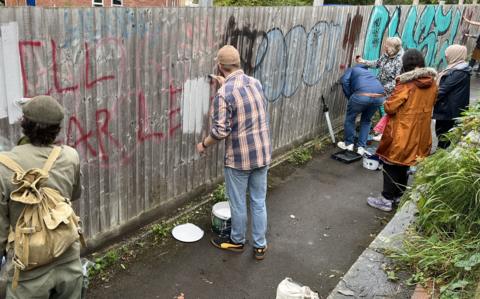 People painting over graffiti on a fence