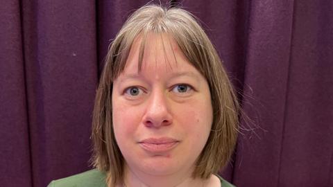 Hilary Parker, a woman with short brown hair wearing a green top and standing in front of a purple curtain