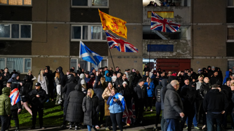Dozens of people, some of them waving Scottish flags, gather outside a block of flats