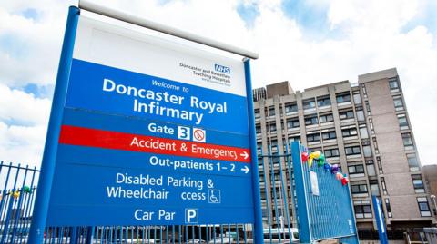 A blue sign at the hospital entrance lists different departments and wards. A car park is behind it, with the words "Welcome to Doncaster Royal Infirmary" written above the building entrance.