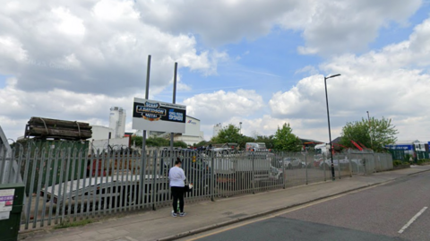 A Google exterior shot of the J Davidson scrap site in Broadheath, Altrincham. The site is behind metal railings. A sign for the firm stands on two poles, and there is a woman walking past the entrance. 