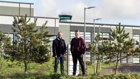 Two men stand in front of an airport terminal with small evergreen trees next to them.