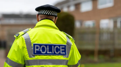 A stock image showing a male police officer wearing a chequered black and white cap and a yellow hi-vis jacket which says 'POLICE' across the back in reflective material. He is facing away from the camera towards a brick building with a tall hedge outside. 