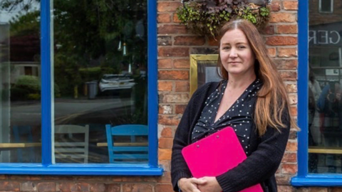 Kelly Woodnutt stood in front of her restaurant St Martha. She is wearing a dark polka dot dress and holding a pink clipboard. Her restaurant is blue.