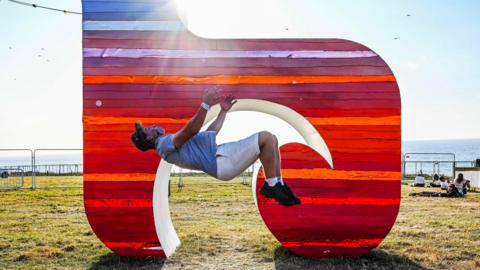 The image shows a person flipping backwards inside a large, colorful sculpture shaped like the letter "b," decorated with red and orange stripes. The person is wearing a gray t-shirt, light shorts, black shoes, and a cap. In the background, there's an open grassy area near the ocean.