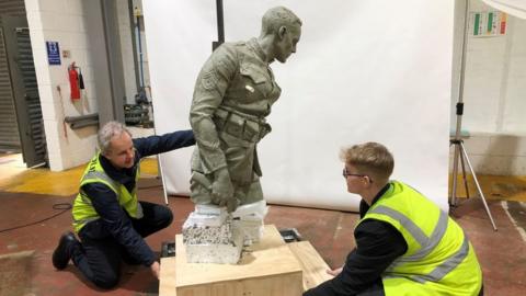Two men holding the wooden base of a sculpture of a WW1 soldier made from clay