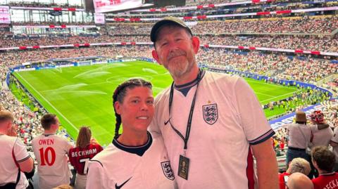 Serena and Dave Stone pose for a picture in a football stadium ahead of England's World Cup match against Croatia. Serena, to the left, has long brown hair. Dave has a beard and is wearing a lanyard around his neck with a pass which has an image of the World Cup on it. There are crowds of people behind them and the football pitch is almost fully visible. Jets of water are being sprayed onto the grass. 