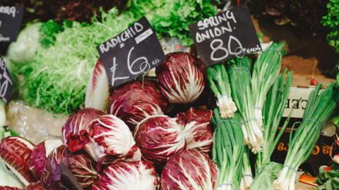 A market stall featuring vegetables and salad, with handwritten black and white price labels. 