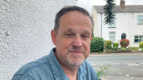 Man in blue shirt outside pebbledash building with a hanging plant and houses behind him 
