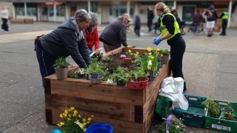 A group of people working together around a large wooden raised planter in an open square, adding soil and arranging colourful flowers. Gardening tools, potted plants and trays are scattered around the planter. In the background, more people are visible near a row of brick buildings with shopfronts and upper‑floor flats. The main workers are three women, and police officer in a high-vis jacket with her blonde hair tied back. 