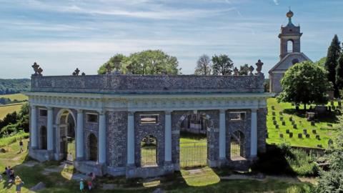 A hexagonal shaped mausoleum on a hill. There is a church and a graveyard behind it. There are people sitting down or walking by it. It is a sunny day with clear blue sky. 
