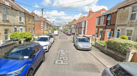 View of a suburban street with cars parked either side of a narrow roadway and clouds in the background.