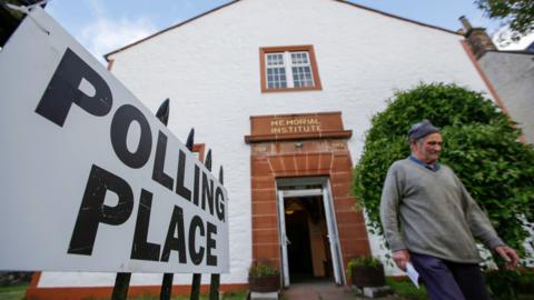 A man wearing a grey jumper and a blue hat leaves after casting his vote at the Moniaive polling station in Dumfries and Galloway. The building has white walls and the doorway is made of red sandstone. A sign on the left of the image says Polling Place.