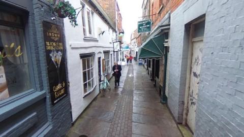 Generic Google shot of a narrow alley with bars and small shops with a brightly coloured bicycle and people in view
