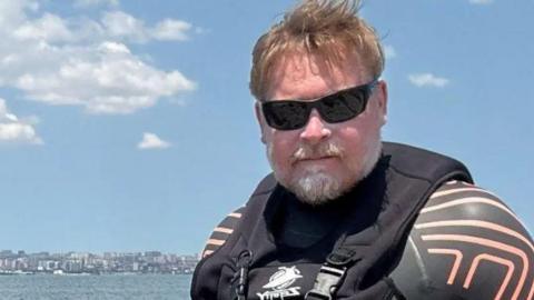 Karl Bushby, wearing sunglasses and a black and red wetsuit, stands on the bank of a calm-looking Caspian Sea. A city can be seen on the far side of the water, while small white clouds float across a blue sky.