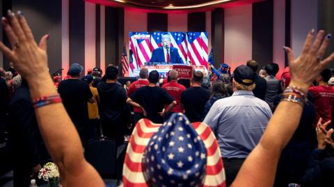 A person wearing a cowboy hat with the US flag on it raises their hands at the back of a room full of people watching Trump's victory speech on a TV screen, at Nevada GOP election watch party in Las Vegas on 6 November