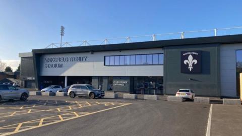 Wakefield Trinity rugby league ground's East Stand at Belle Vue, with blue skies above and a car park in front