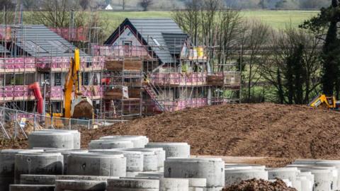 A building site with green fields in the background. Several new builds can be seen with scaffolding around them.