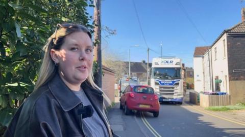 A woman looks concerned as she stands on a footpath by the side of a narrow residential road as a red car mounts the pavement to avoid white HGV lorry heading in the other direction. The woman has dark-blond hair and wears a black leather jacket, grey-blue T-shirt and sunglasses on the top of her head. Green shrubs and white-washed houses can be seen. The sky is blue.