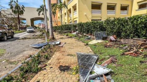 A yellow building with palm trees outside is littered with broken solar panels and ripped up shrubs