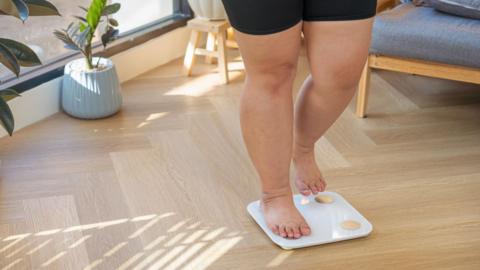 Waist down shot of a woman standing on weighing scales, wearing black gym shorts. She has bare legs and feet.  She is in a bright living room which has a wooden floor and potted plants in the background. 