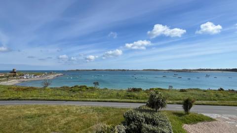 The image shows Grand Havre bay. There is a road and behind it is a stretch of green grass and then blue sea with moored boats. The sky is blue with white clouds.