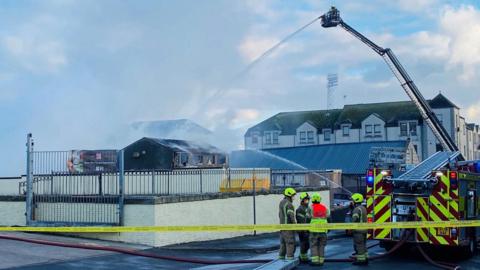 Fire engine and firefighters, with plume of grey smoke, and a height vehicle being used to dampen down the scene, with football stadium floodlights in the background.