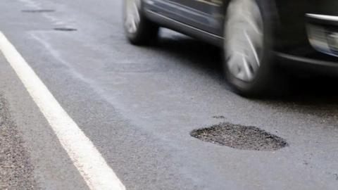 A close up image of a pothole on a road with a car wheels driving close to it