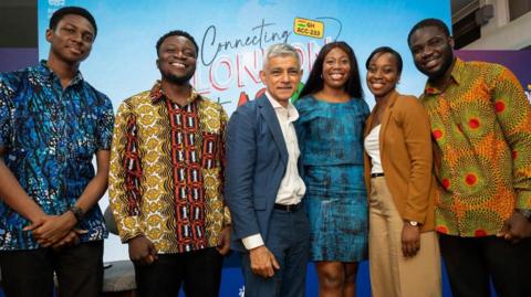 Sadiq Khan stands at the centre of a group of people posing for a photo. He is wearing a blue suit and white shirt. 