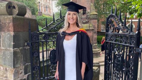 A woman with blonde hair wearing a black mortarboard and a black graduation robe with red and yellow near the neck. She is standing in front of small black iron gates.