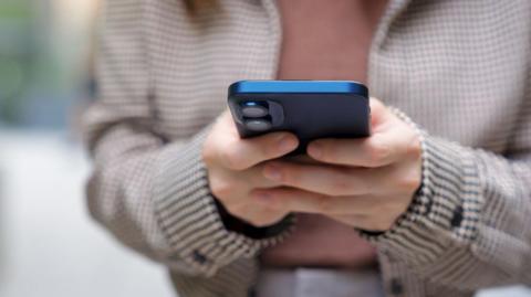 Hands holding a blue smartphone outdoors. The person wears a checkered coat and a pink top, and the background is softly blurred.