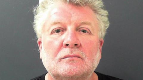 Close up shop of a man with a short white beard and white hair. He has grey eyes and is looking directly into the lens with a neutral, slightly sad expression.