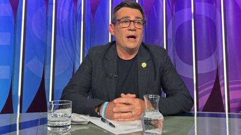 Andy Burnham is sat down, with a glass of water and a jug of water in front of him. He is wearing a dark coloured jacket, a dark coloured top and glasses.
