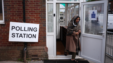 A polling both on a residential street, with a large white sign next to the door. A woman in a brown headscarf is leaving the building.