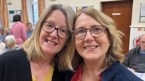 Two women standing next to each other with their heads touching. The woman on the left has shoulder-length blonde hair and is wearing black-framed glasses, a mustard yellow T-shirt and a black jacket. The woman on the right has shoulder-length blonde hair and is wearing brown-framed glasses, a pink patterned T-shirt and a navy jacket. People are sitting behind them on chairs.