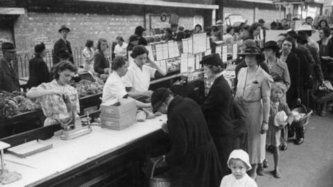 Shoppers queuing to buy food at a stall run by a department store at an open air market at Plymouth in the wake of German bombing during World War Two