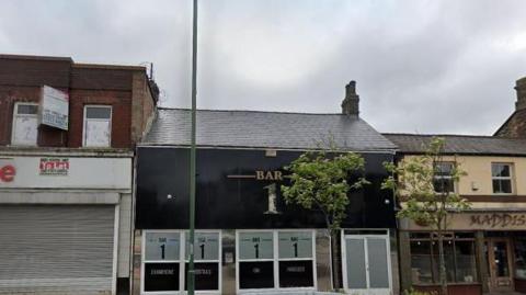 The front of Bar1 in Consett. A black fronted building with four windows and a door on the ground level and a large sign above containing the bar's name.