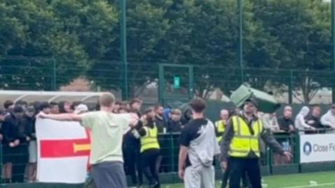 Football fans stood on a pitch. Some are stood behind a gate. There is a bin in the air and trees in the background.
