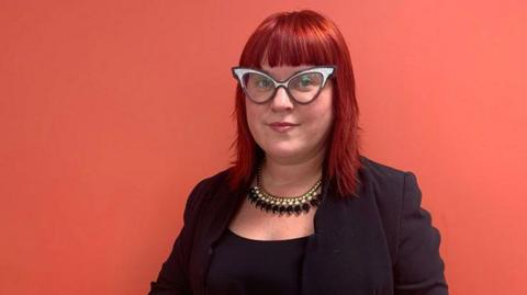 Dr Sally Pezaro wearing a very large pair of white and brown framed glasses,  in a black shirt and jacket standing against a salmon pink background