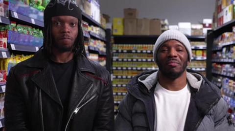 Krept and Konan are seen standing side-by-side facing the camera in an aisle of their supermarket.