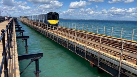 A picture of a Network Rail train, which has yellow detailing at the front and black panelling, is riding along a railway line, fixed above the sea on the Island Line. A pier runs alongside the line, decorated with black metal railings and people can be seen walking along the pier in the distance.