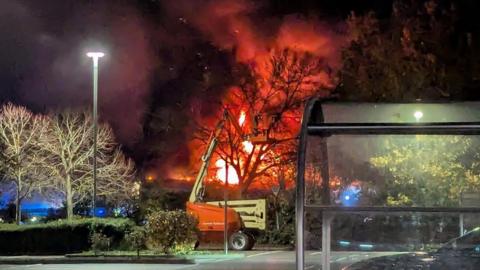 A bright fire lights up a plume of smoke against a dark sky. The fire is taking place behind an orange cherry picker near a tree and a hedge. The picture is taken from an otherwise empty car park.