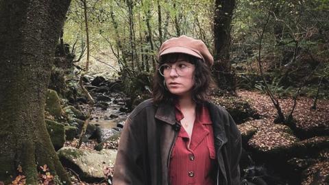Alice, a woman with shoulder-length brown hair, is wearing a beige cap and large glasses. She looks to one side and is standing in a glen, with a shallow stream behind her.