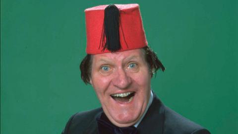 Tommy Cooper is in the middle of the picture, smiling. The picture is taken from above the shoulders, and he is wearing a black suit jacket, and bow tie. He is wearing a fez, which is a red hat with a black tassel on. 